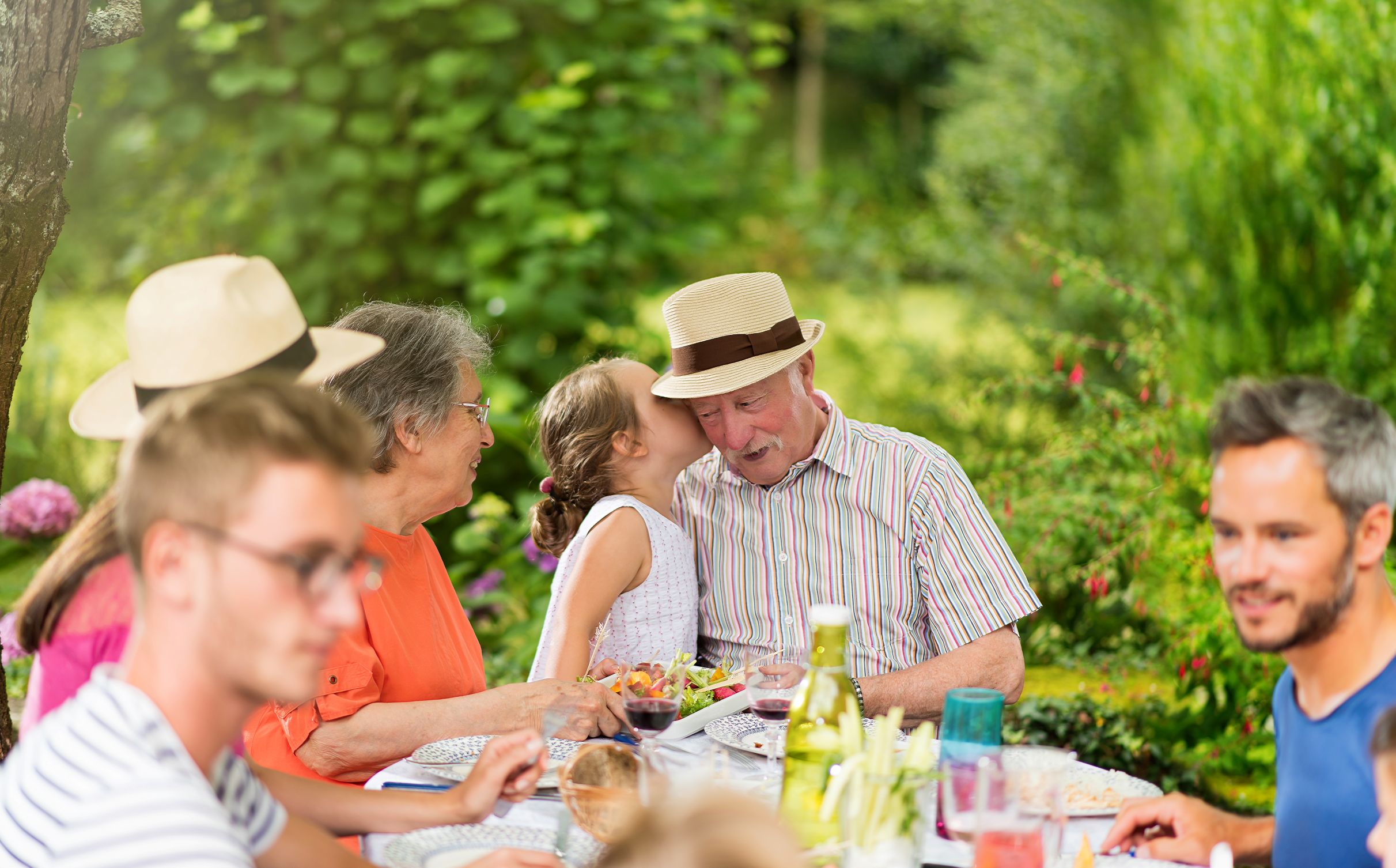 Familie sitzt an einem Gartentisch mit Picknick, umgeben von grüner Natur, Kinder und Erwachsene genießen das Essen