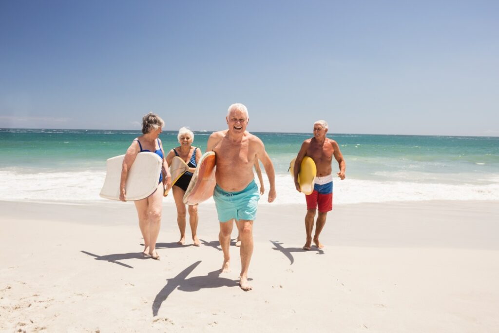 Fünf ältere Menschen mit Surfboards laufen barfuß am Strand entlang, Meer und blauer Himmel im Hintergrund
