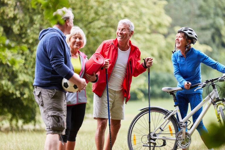 Vier ältere Menschen im Park, eine Frau mit Fahrradhelm und Fahrrad, ein Mann mit Wanderstöcken, eine Frau hält eine Yogamatte und ein Mann hält einen Fußball