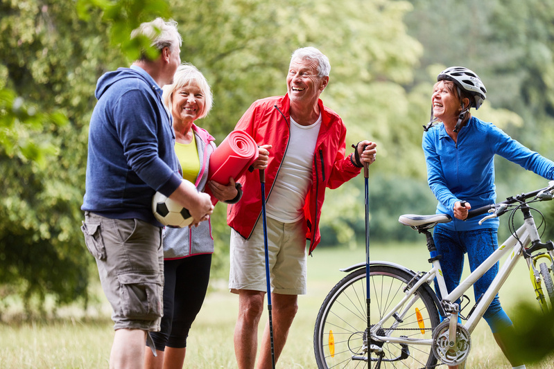Vier ältere Menschen im Park, eine Frau mit Fahrradhelm und Fahrrad, ein Mann mit Wanderstöcken, eine Frau hält eine Yogamatte und ein Mann hält einen Fußball
