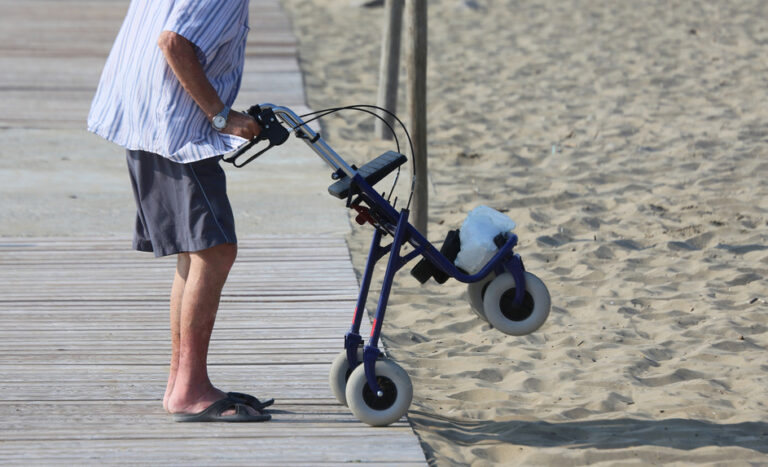 Älterer Mann mit Rollator steht auf Holzsteg am Strand