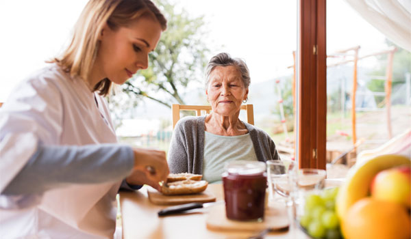 Eine 24-Stunden-Betreuung schmiert für eine Seniorin ein Brötchen.