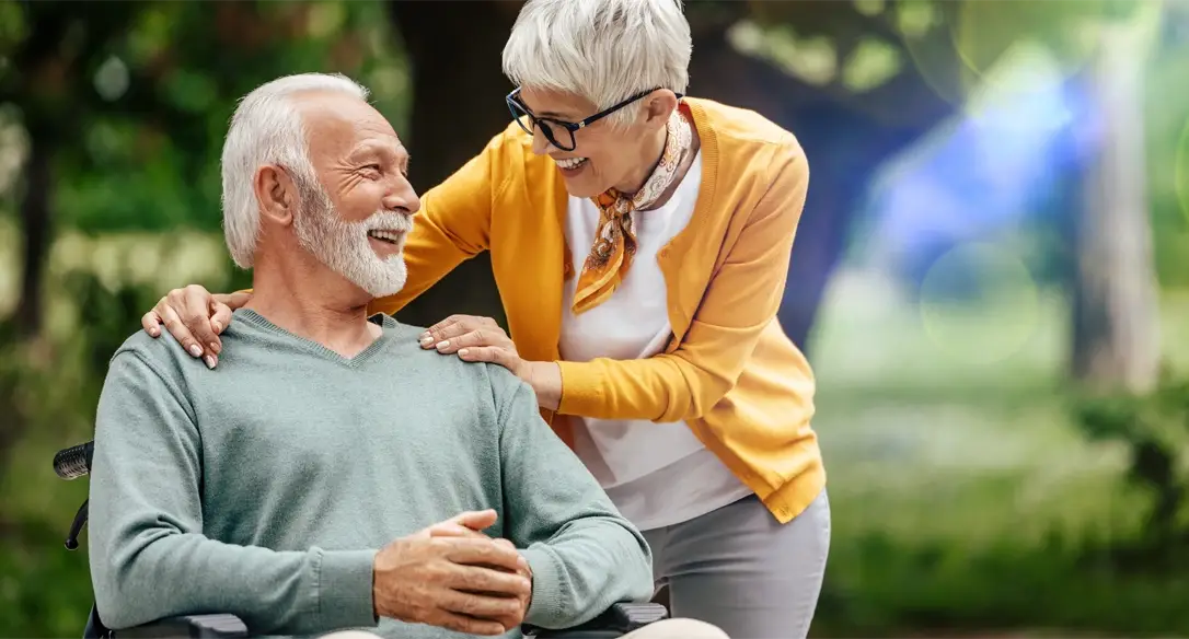 Ältere Person mit grauem Pullover sitzt, während eine andere Person mit orangefarbenem Pullover sie von hinten umarmt.