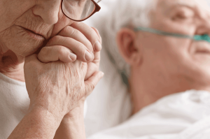 Eine Seniorin hält die Hand ihres Mannes, der schwer krank im Bett liegt.