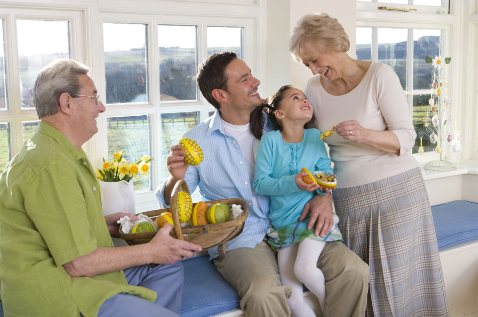 Oma überreicht Enkelin ein Schokoladenosterei, während die Enkelin auf dem Schoß des Vaters sitzt.