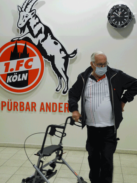 Ein Mann mit Rollator steht vor dem FC-Wappen.