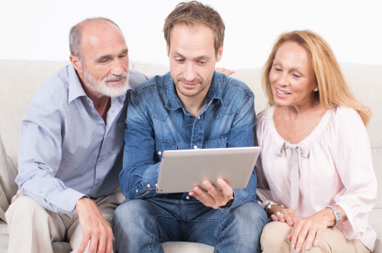 Sohn sitzt mit Eltern auf der Couch. In seiner hand ein Tablett mit dem er die Kosten für einen Hausnotruf berechnet.