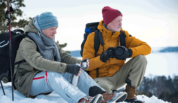 Vater und Sohn machen im Winter eine Rast. Der Vater hat eine Kamera dabei und ist auf Motivsuche. Der Sohn gießt sich einen heißen Tee ein. 