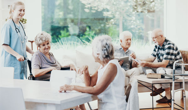 Eine Pflegerin schiebt eine Seniorin mit dem Rollstuhl ins Wohnzimmer einer Senioren WG.