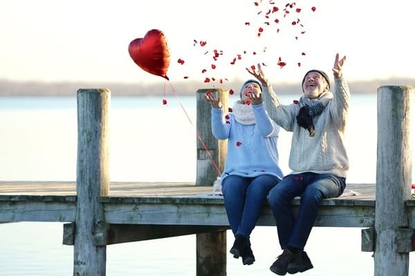 Ein Rentner Ehepaar sitzt an einem Holzsteg. Er schmeißt Rosenblätter in die Luft, sie hat einen roten Herz-Luftballon in der Hand