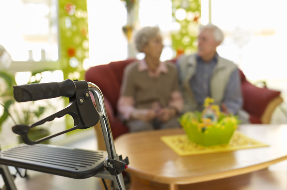 Rollator im Vordergrund. Ein Seniorenpaar auf der Couch im Hintergrund