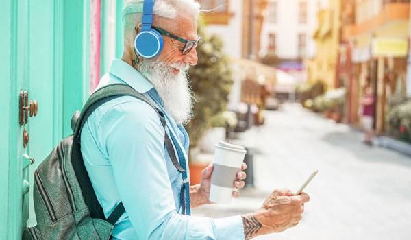 Senior mit Hippster Bart, Rucksack und Kopfhörer schaut auf sein handy. Dabei steht er in einer Straße mit bunten Häusern.