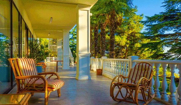 Eine Seniorenresidenz im Ausland mit weitläufigen Balkon und Korbmöbel zum sitzen. Dabei in einen grünen Garten mit Tanne und einem See blicken.