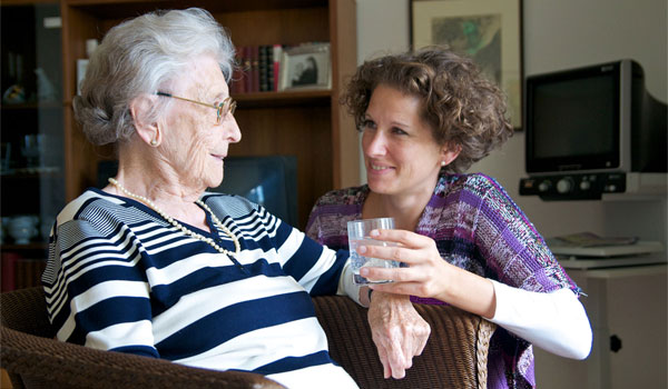 Tochter reicht ihrer alten Mutter, welche im Sessel sitzt, ein Glas Wasser.