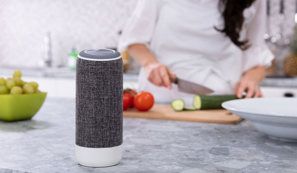 Ein Sprachassistent steht auf einer Arbeitsplatte in der Küche. Während eine Frau Gurken schneidet.