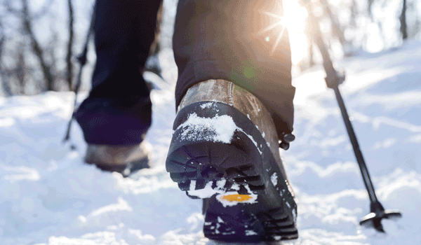 Eine Person geht mit Skistöcken durch den Schnee. Dabei sind nur die Wanderschuhe mit Profil und dessen Beine zu sehen.