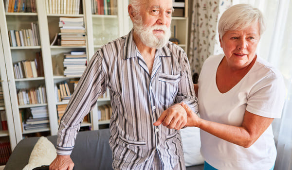 Ein Senior mit einem Schlafanzug bekleidet, wird von seiner Frau gestützt.