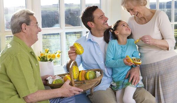 Oma überreicht Enkelin ein Schokoladenosterei, während die Enkelin auf dem Schoß des Vaters sitzt.