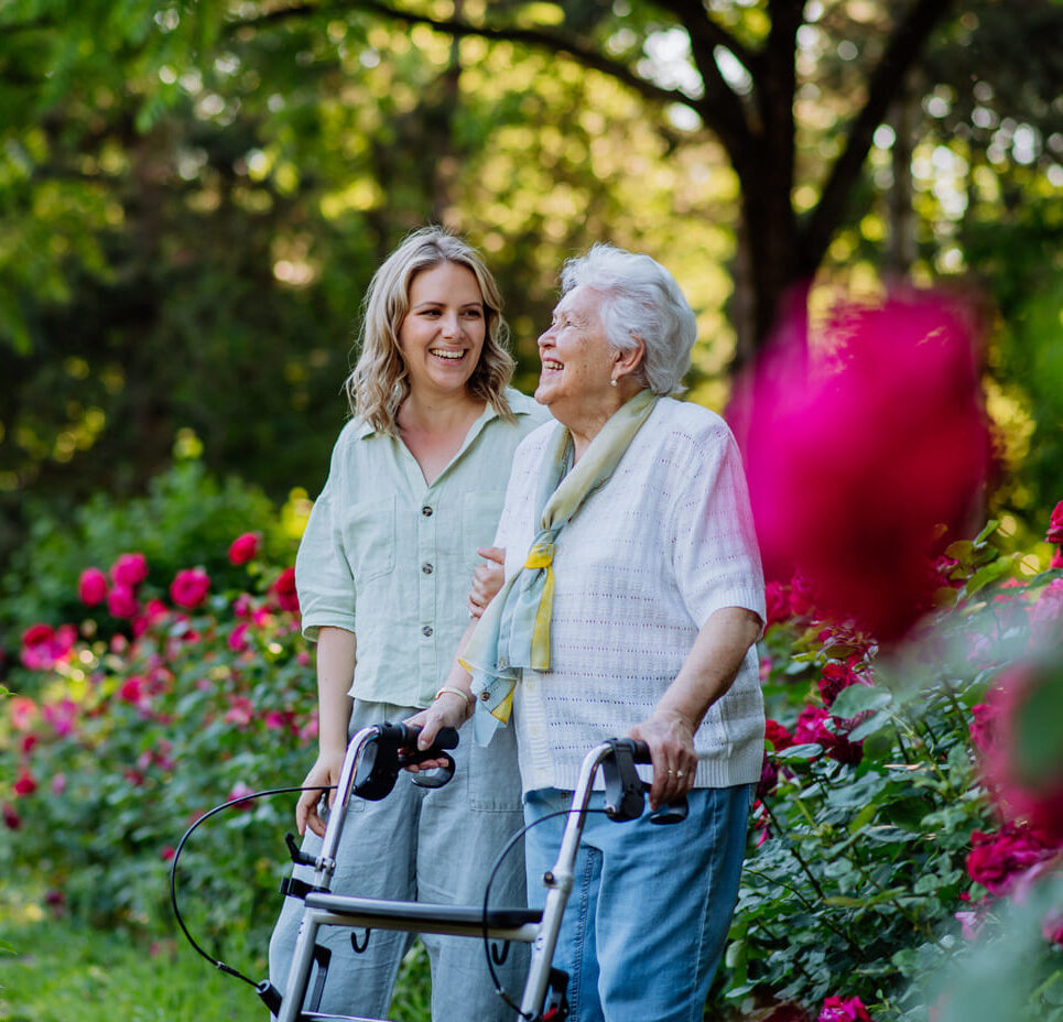 Eine ältere Frau mit einem Rollator geht mit einer Freundin im Garten spazieren