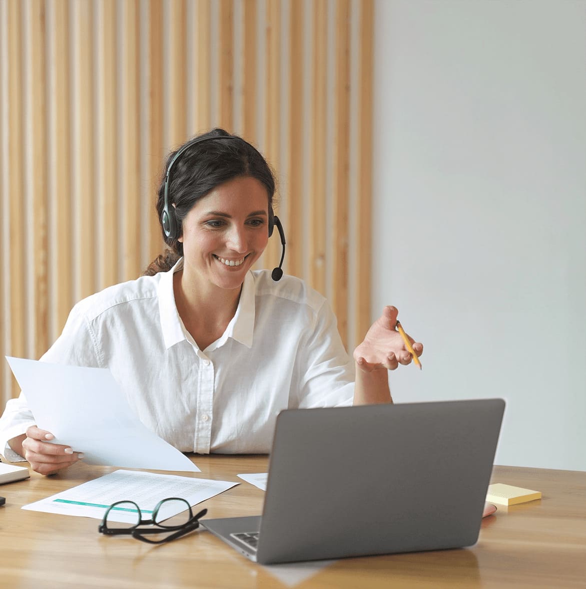 Eine Frau mit weißem Hemd und Headset sitzt an einem Holztisch vor einem Laptop, hält Papier und einen Bleistift, auf dem Tisch liegen Brille und weitere Papiere.