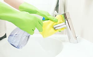 Person mit grünen Handschuhen reinigt Wasserhahn mit gelbem Tuch und Sprühflasche