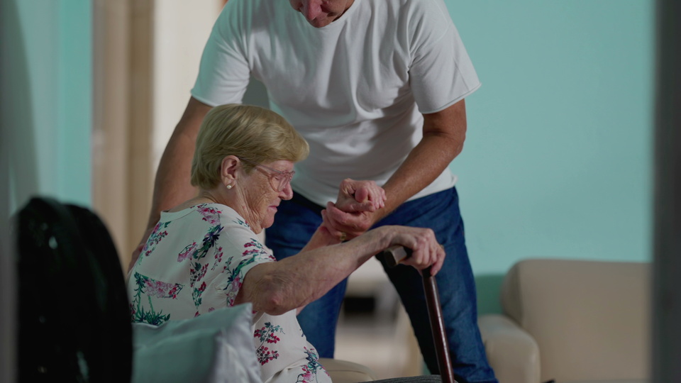 Ein Mann hilft einer älteren Frau beim Aufstehen. Sie hält einen Stock in der Hand.