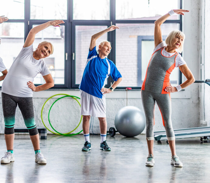 Drei ältere Menschen in Sportkleidung machen seitliche Dehnübungen in einem hellen Fitnessraum mit großen Fenstern