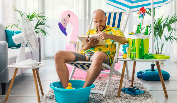 Ein Mann sitzt zu Hause im Strandstuhl. Seine Füße baden in einer Schüssel, links neben ihm ein Ventilator und rechts Erfrischungsgetränke.