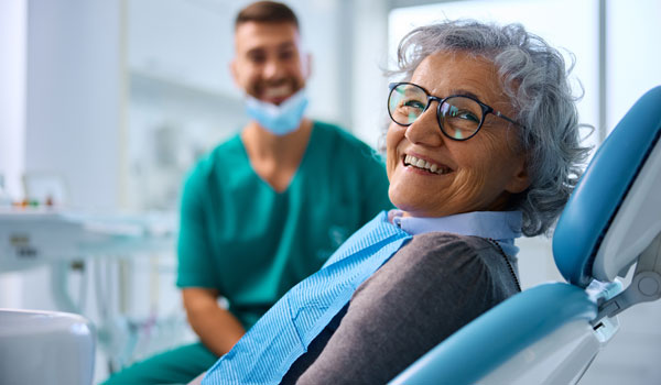 Eine Seniorin sitzt beim Zahnarzt auf dem Behandlungsstuhl. Im Hintergrund ist der Arzt zu sehen.