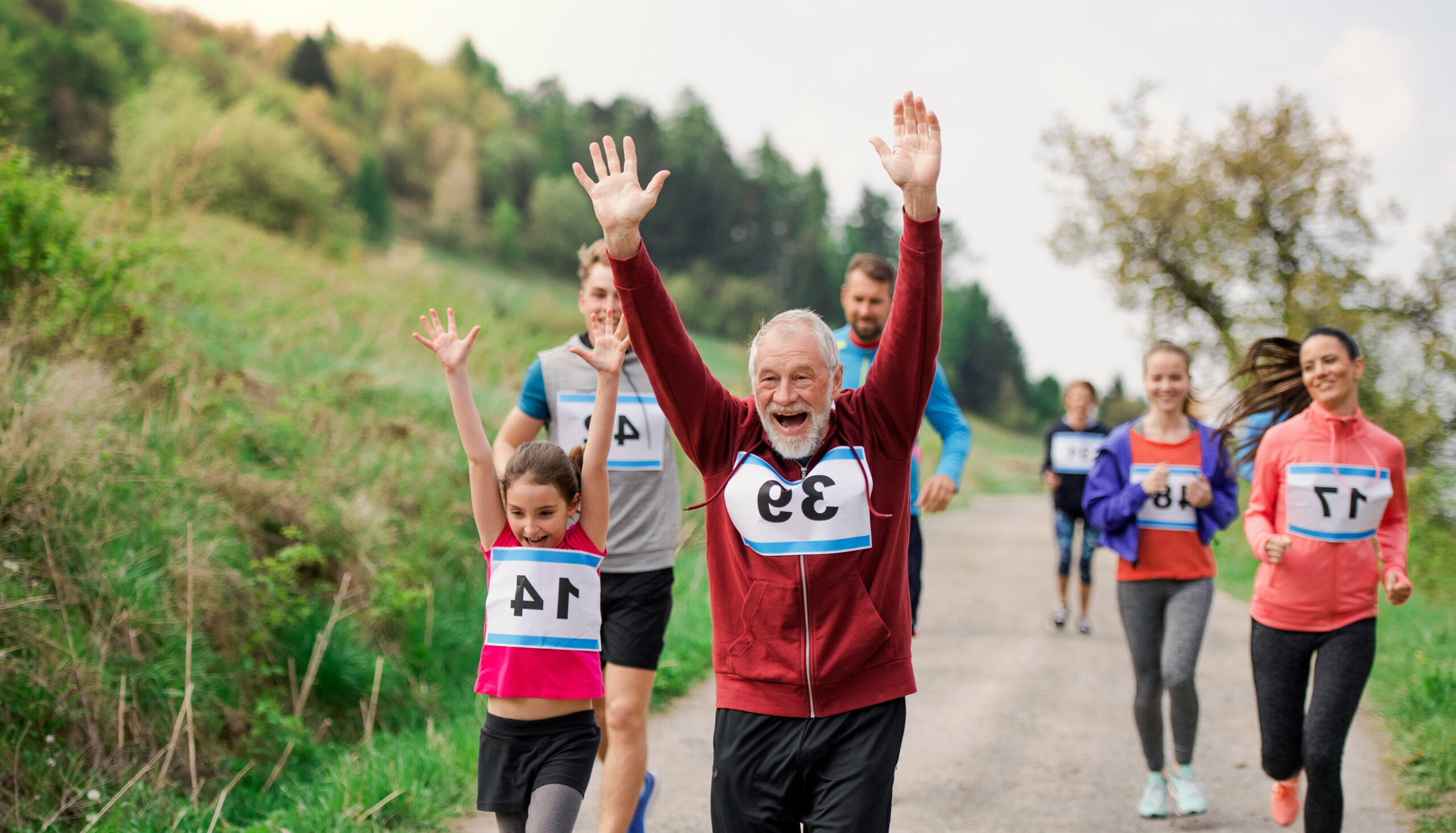 Mehrere Läufer mit Startnummern auf einem Waldweg, zwei vorne mit erhobenen Armen, Teilnehmer verschiedener Altersgruppen