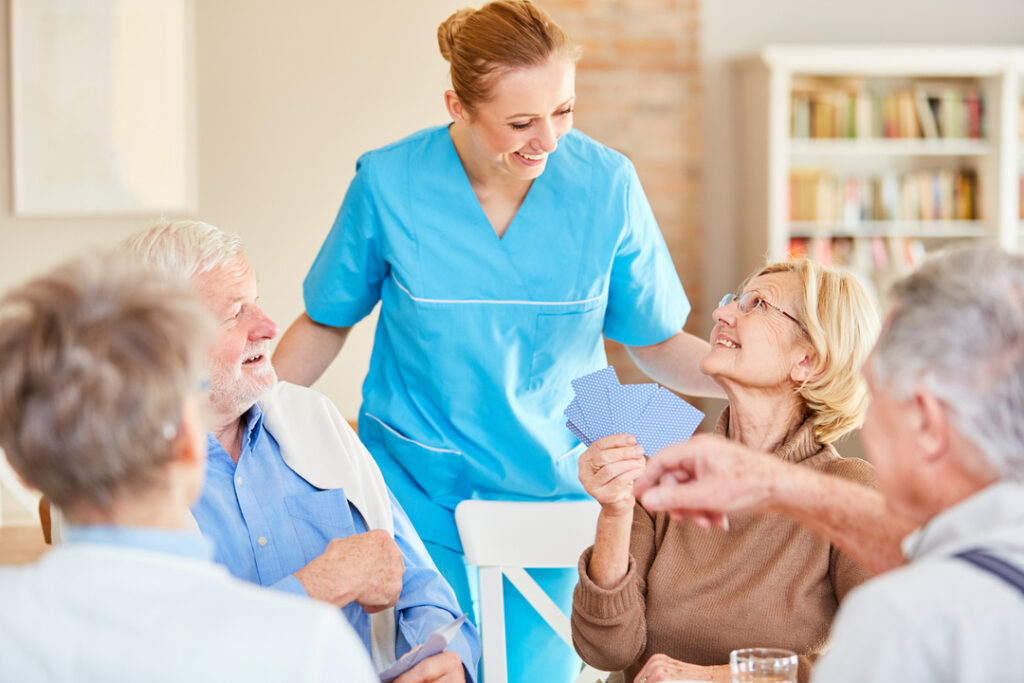 Pflegekraft in blauer Uniform steht am Tisch mit drei älteren Menschen, die Karten spielen