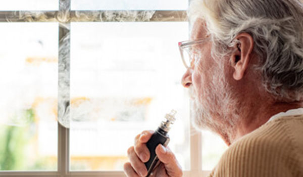 Ein älterer Mann steht vor seinem Fenster und raucht dabei eine E-Zigarette.