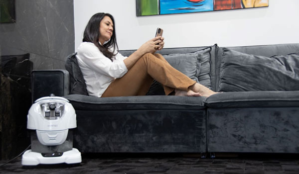 Eine Frau sitzt auf ihrem Sofa und liest auf ihrem Handy, während ihr Staubsauger vor der Couch steht.