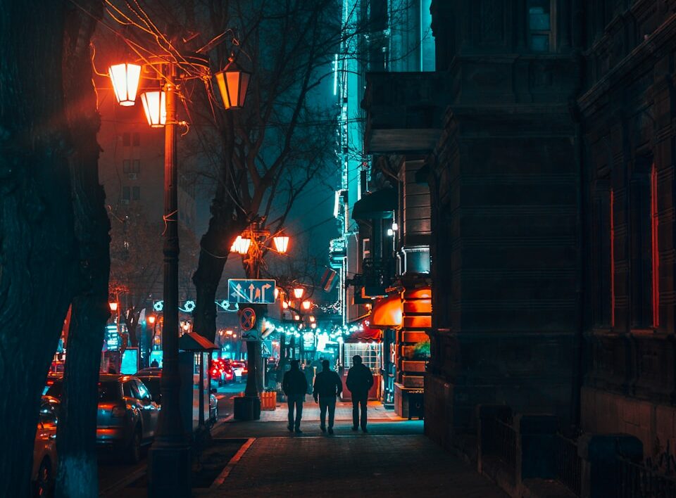 Drei Personen gehen nachts auf einem beleuchteten Bürgersteig einer Stadtstraße mit Straßenlaternen und Autos