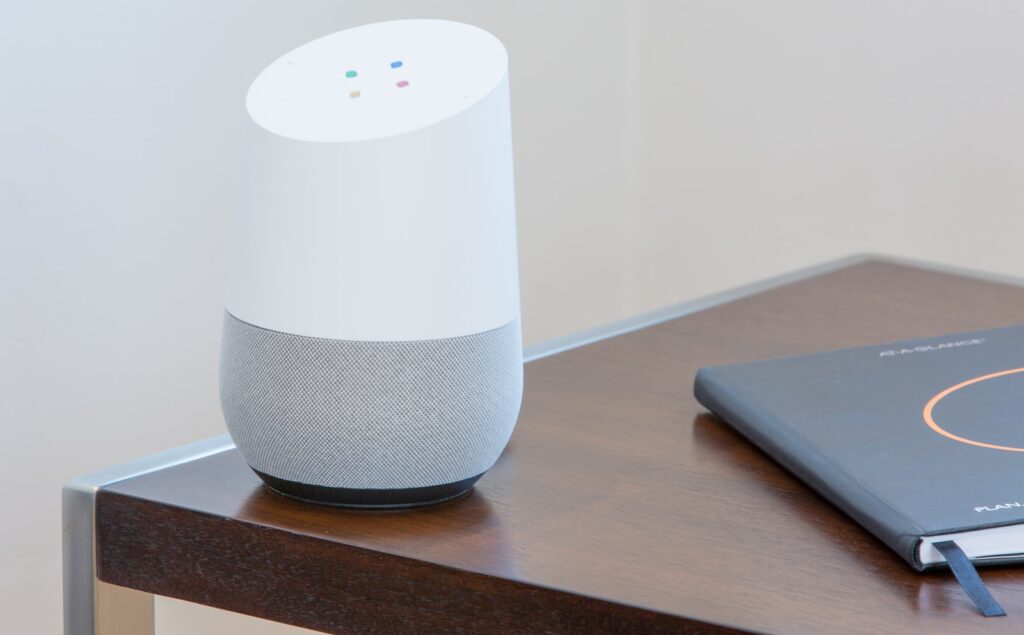 Weißer Google Home Lautsprecher auf einem dunklen Holztisch neben einem geschlossenen Notizbuch