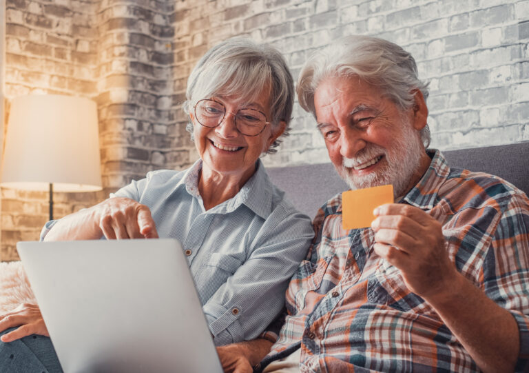 Porträt eines Paares von zwei niedlichen, glücklichen und aufgeregten Senioren, die mit ihrem Laptop zu Hause auf dem Sofa sitzen und online einkaufen und Geld ausgeben. Sie kaufen gemeinsam mit ihrer Kreditkarte im Internet ein.