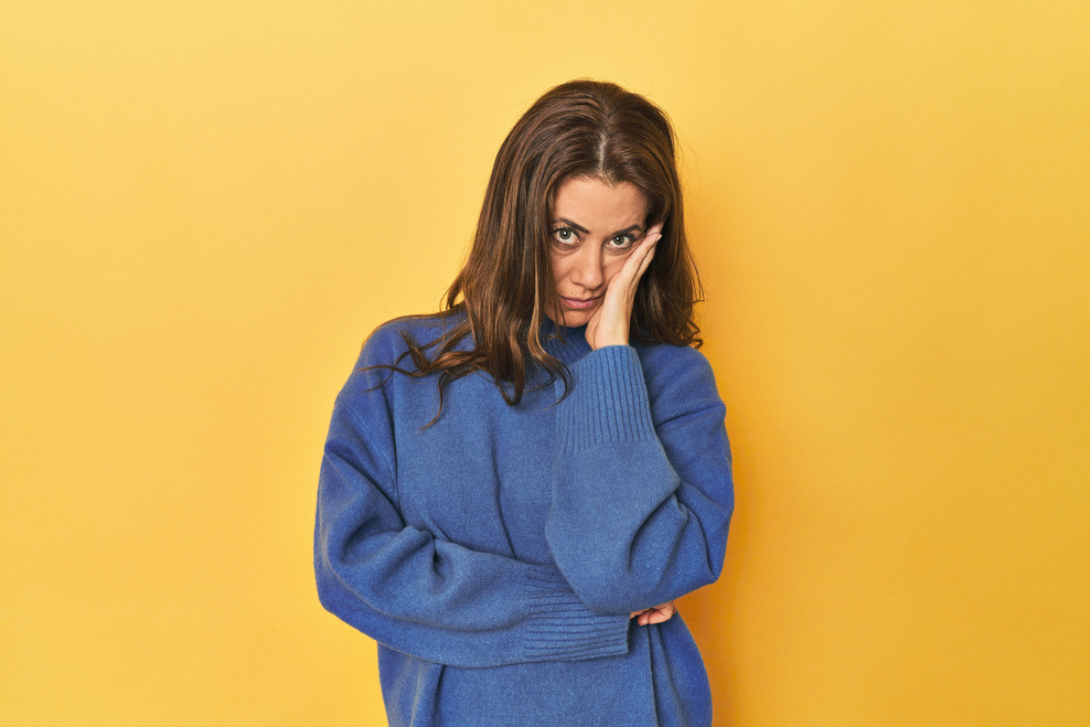 Eine pflegende Angehörige mittleren Alters vor einem gelben Hintergrund. Sie sieht erschöpft aus und braucht eine Pause.