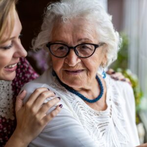 Portrait einer alten Frau, die von einer jüngeren an den Schultern gehalten wird.