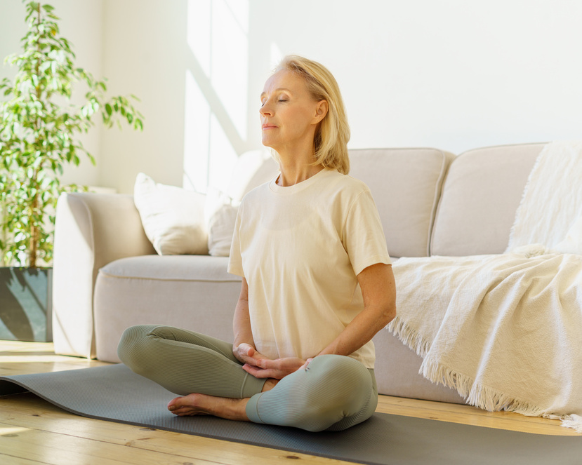 Eine Seniorin sitzt im Schneidersitz vor ihrer Couch und macht Atemübungen.