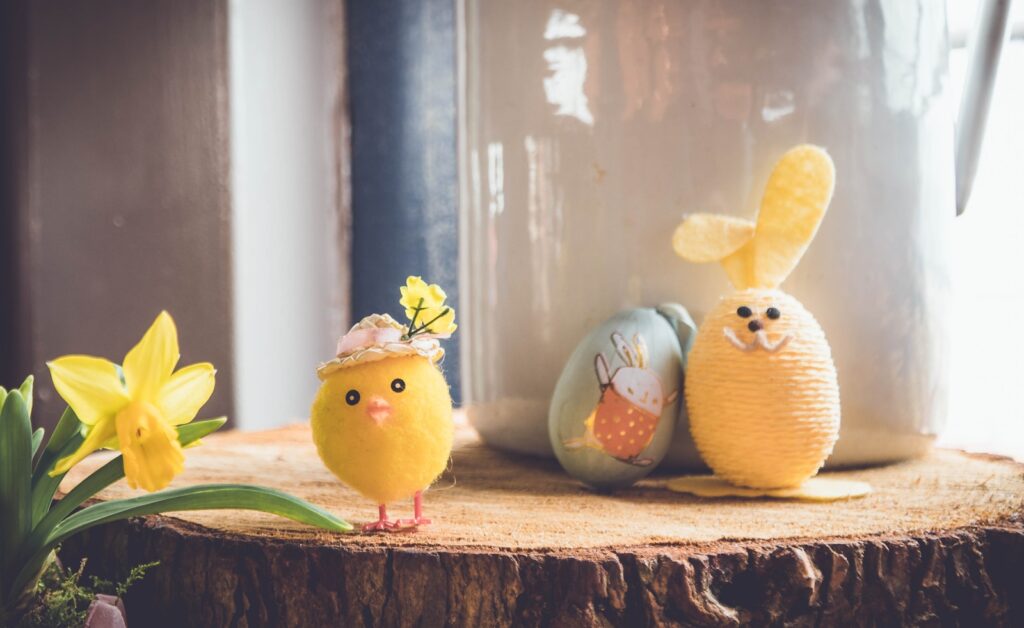 Gelbes Küken mit Blumenhut, bemaltes Osterei mit Hasenmotiv und gelber Osterhase aus Garn auf Holzscheibe neben Narzisse